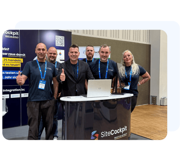 Six members of the SiteCockpit team stand at a trade fair booth behind a counter with a laptop and present the solution at a trade fair
